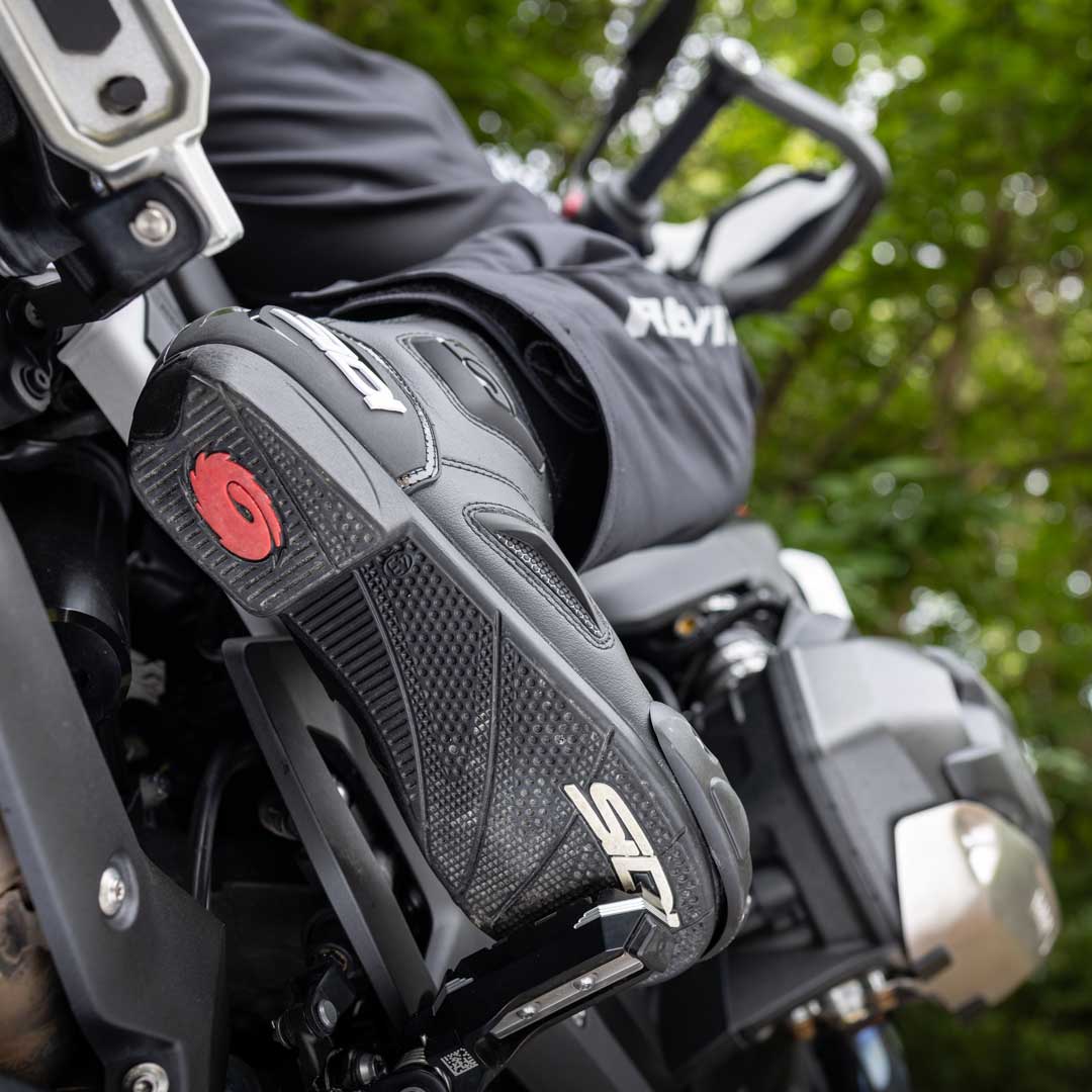 Close-up of a person wearing black motorcycle boots with red accents on a motorcycle, with a blurred green background.